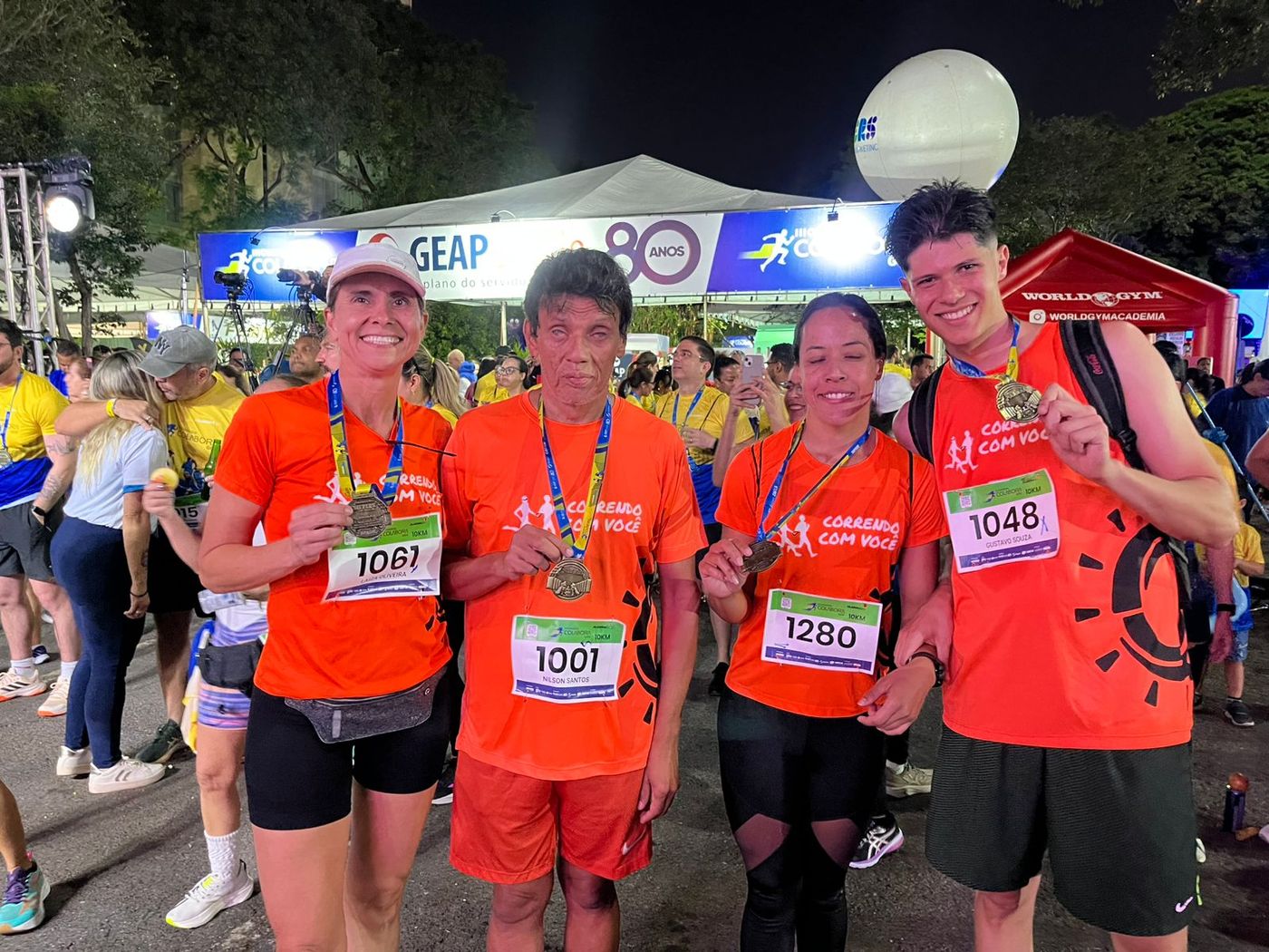 Quatro corredores comemorando à noite com as medalhas de participação após uma corrida de 10 km, em frente a um palco e uma faixa “GEAP 80 anos”.