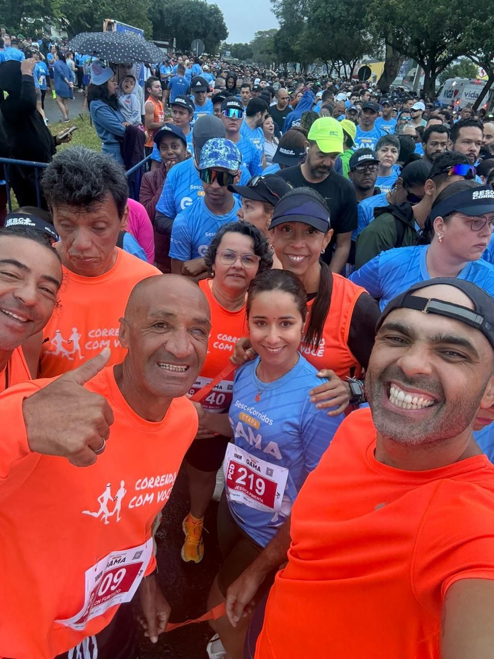 Foto de perto do grupo de corredores com camisetas laranjas, cercados por uma grande multidão de participantes com camisetas azuis na linha de largada.