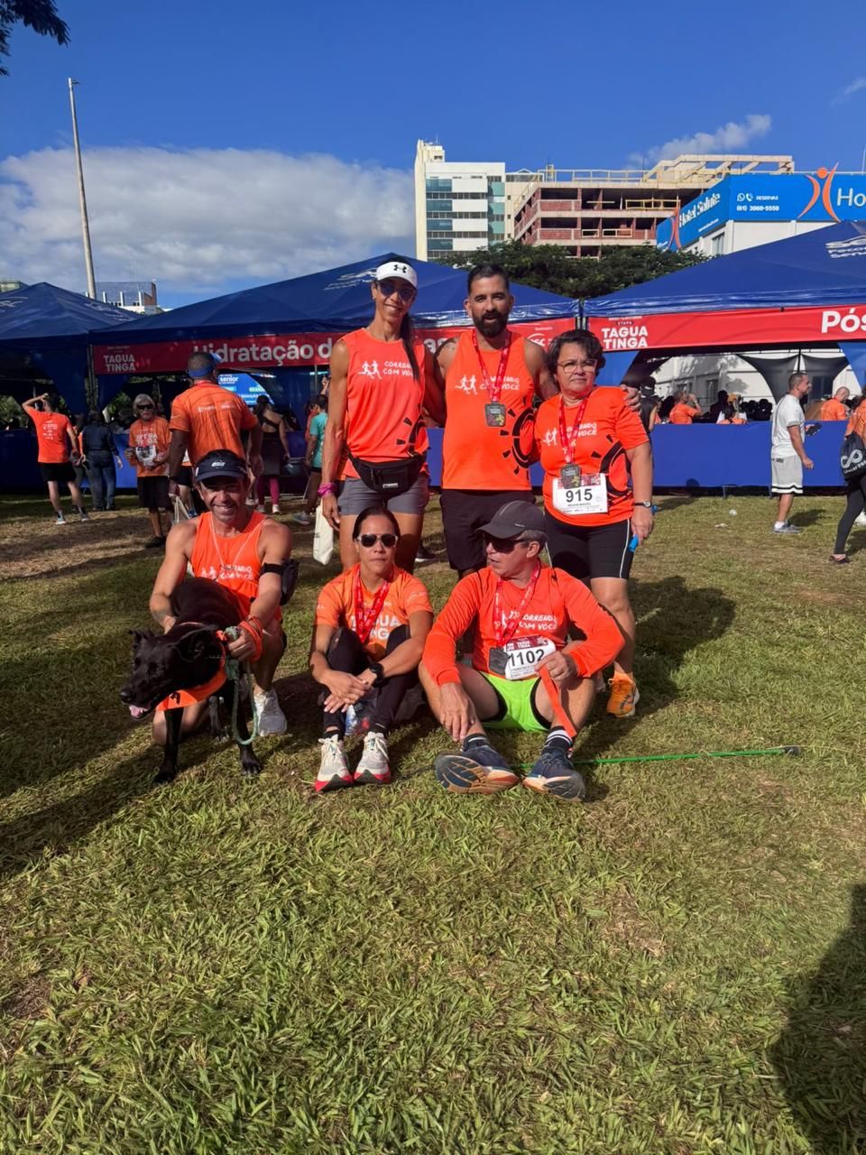 Corredores relaxando na grama após uma corrida, com um cachorro usando uma bandana laranja.
