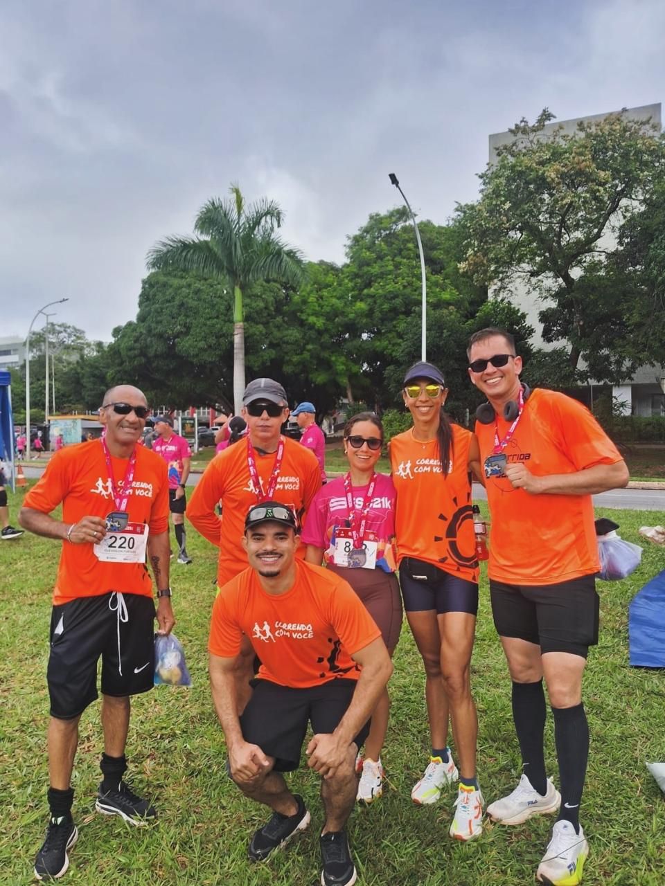 Seis corredores posando juntos com suas medalhas após uma corrida, com uma palmeira e céu nublado ao fundo.