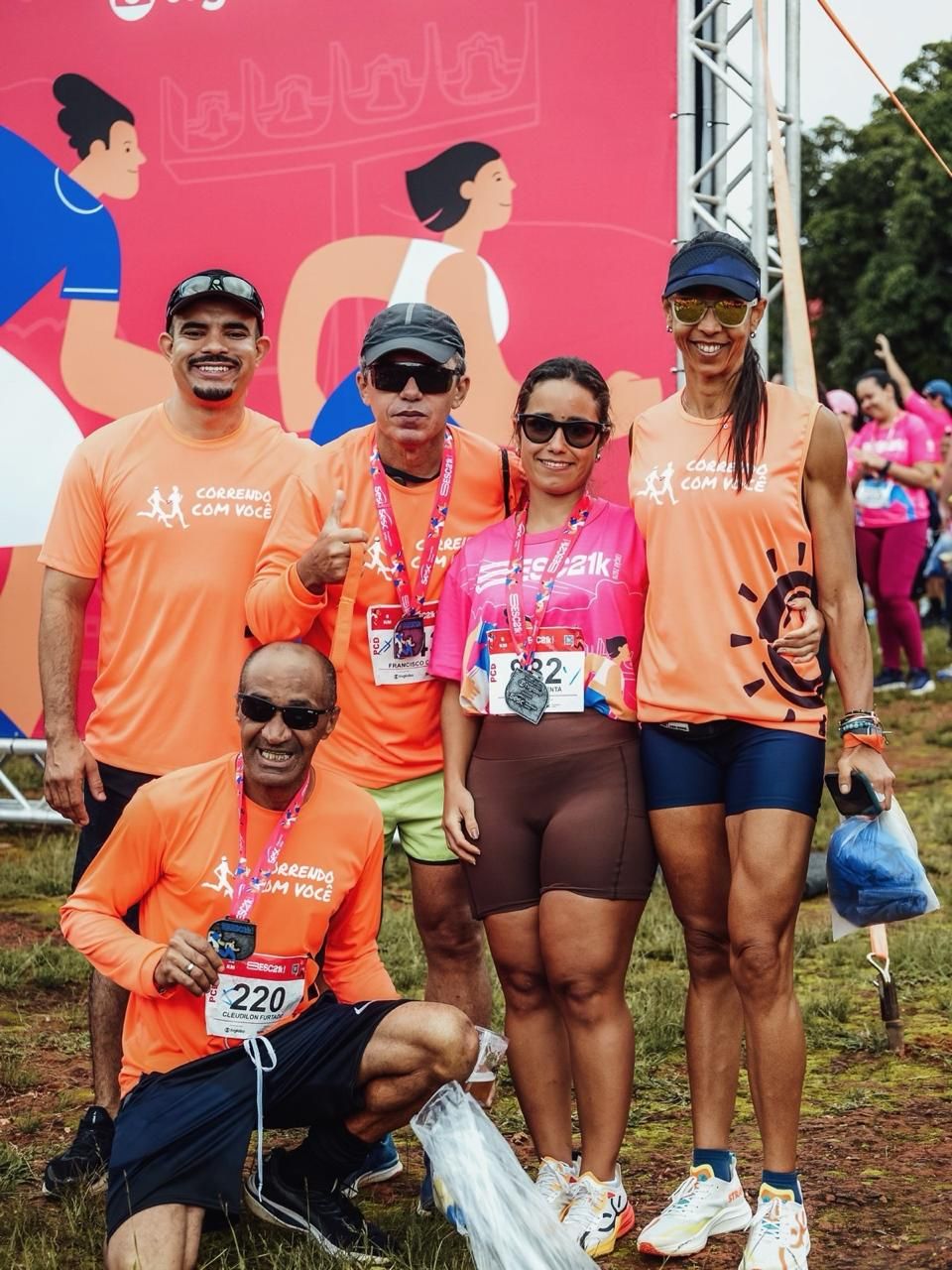 Cinco corredores posando juntos com suas medalhas em frente a uma faixa rosa da corrida.