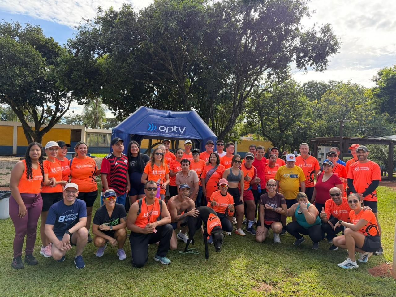 Grande grupo de integrantes do Correndo Com Você reunidos perto de uma tenda inflável azul, incluindo um cão-guia usando um colete laranja.