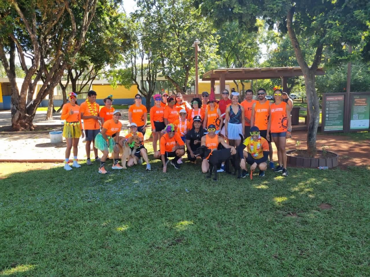 Grupo de corredores comemorando fantasiados, usando colares de flores, perucas e óculos coloridos, com dois cachorros, durante uma corrida temática festiva.