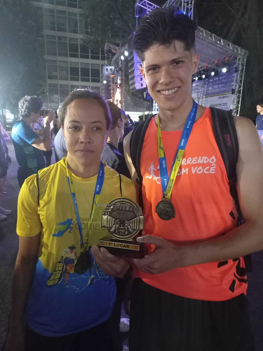 Um jovem com uma regata laranja do Correndo Com Você e uma mulher com uma camiseta amarela de corrida sorriem enquanto seguram juntos um troféu de primeiro lugar, ambos usando medalhas, em uma corrida noturna com um palco iluminado ao fundo.