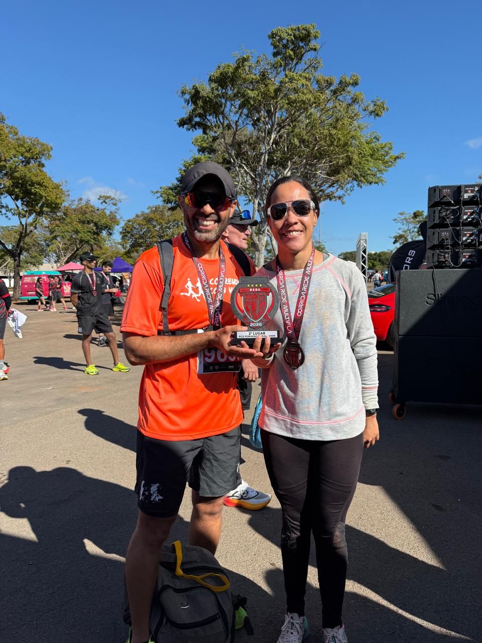 Um homem com uma camiseta laranja do Correndo Com Você e uma mulher com um moletom cinza claro sorriem enquanto seguram juntos um troféu de segundo lugar ao ar livre em um dia ensolarado.