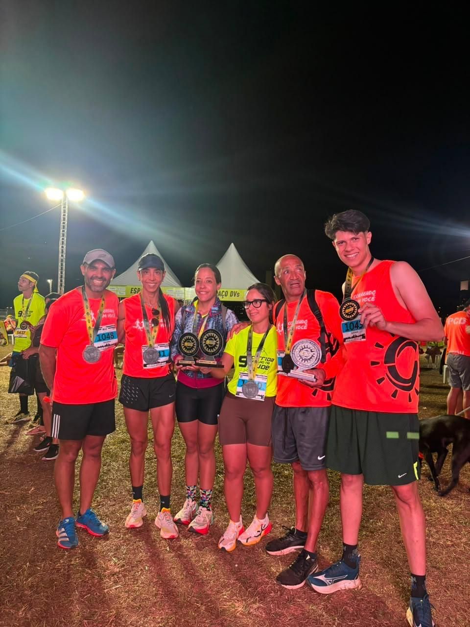 Seis integrantes do Correndo Com Você posando à noite com medalhas e troféus após uma corrida, com barracas ao fundo e um cachorro por perto.