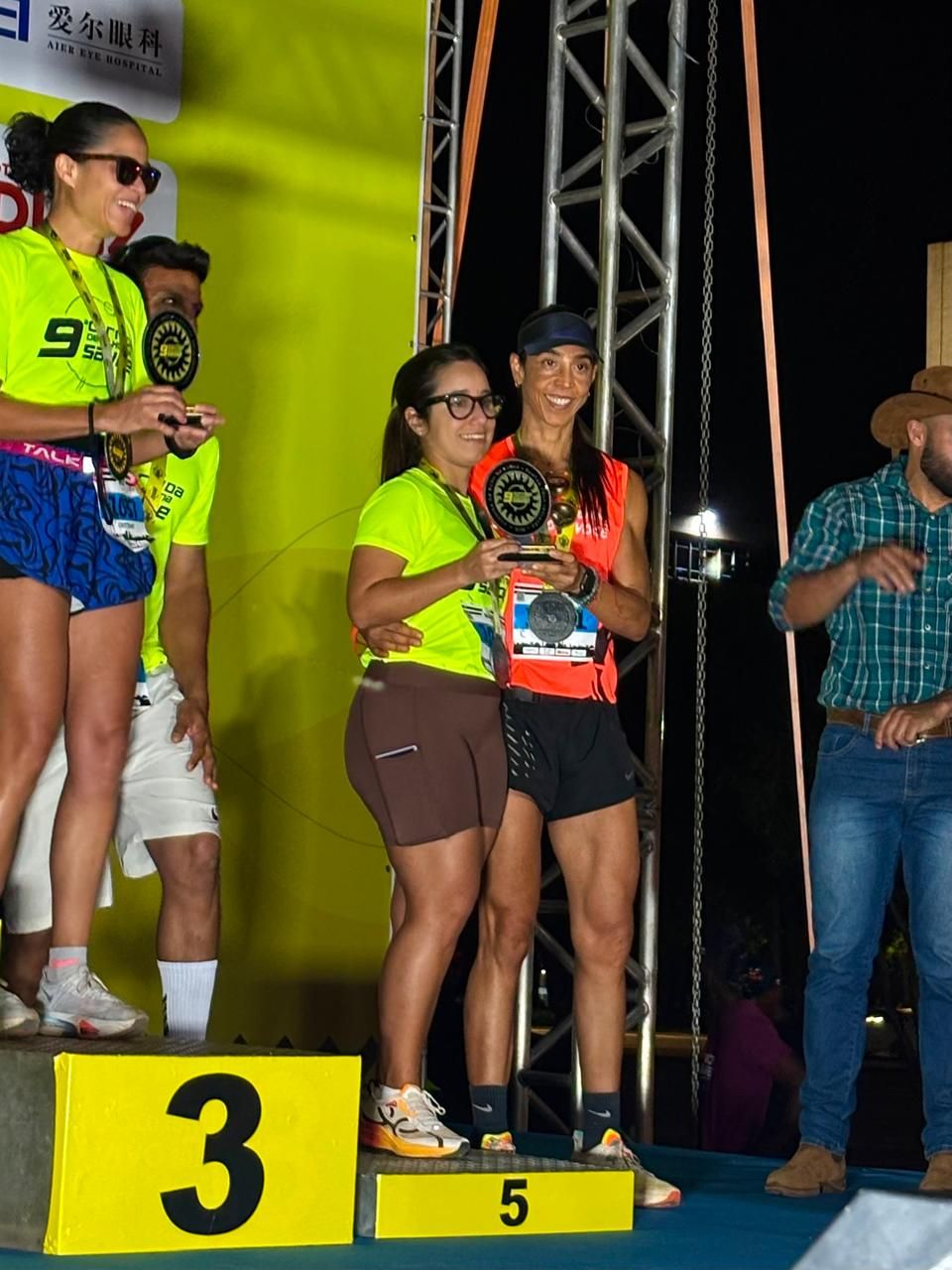 Duas corredoras se abraçando e sorrindo em um pódio, uma com camiseta amarelo-neon e outra com regata laranja, segurando um troféu, no degrau número 3.